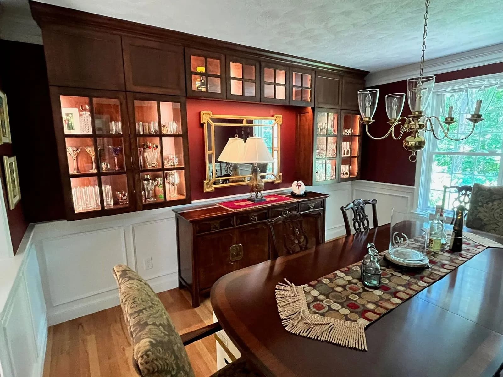 A wide view of a formal dining room with lit glass-front built-in cabinets, a red accent wall with gilt mirror, wainscoting and a brass chandelier.