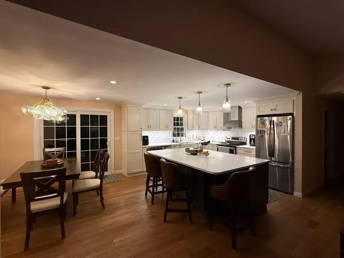 A remodeled kitchen at night with three glass pendants over a white island, recessed lights and under-cabinet lighting, and a bubble-glass chandelier over the dining table by the patio doors.