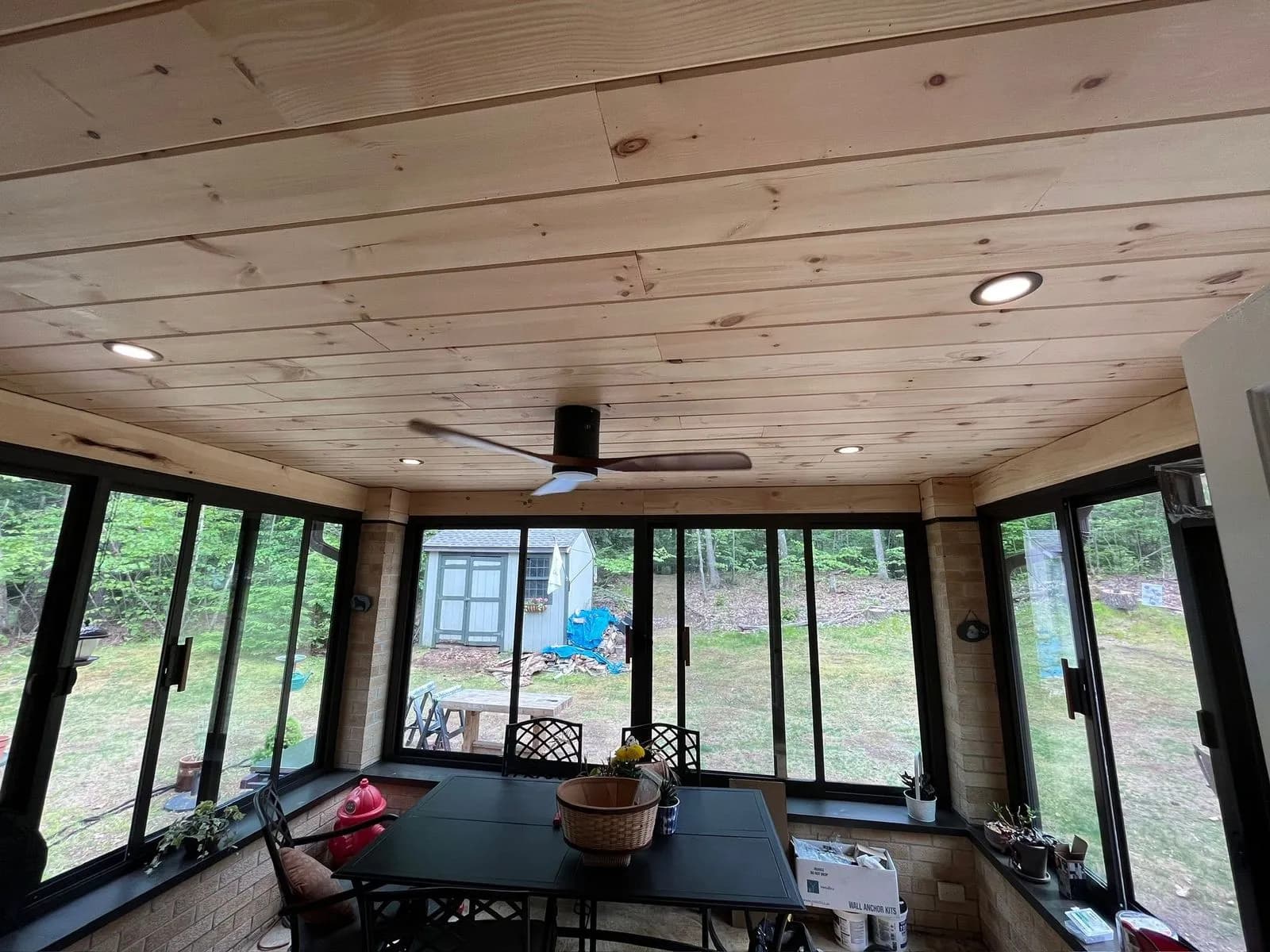 A three-season sunroom with a knotty pine ceiling, recessed lights and a black modern ceiling fan, windows all around looking out on a backyard and shed.