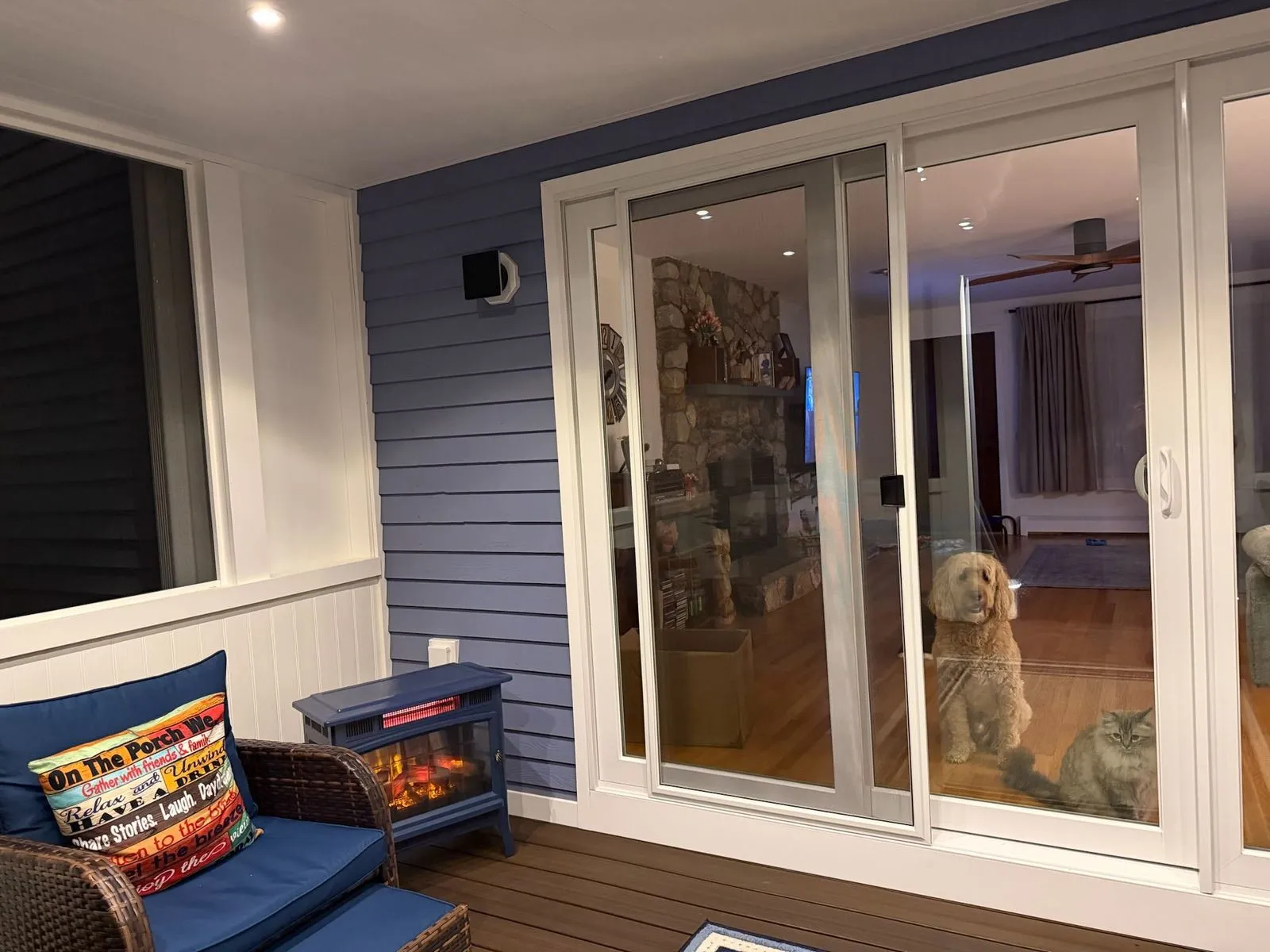 A screened porch at night lit by recessed ceiling lights, with blue wicker seating, an electric fireplace heater and a dog and a cat looking out through the sliding glass door.