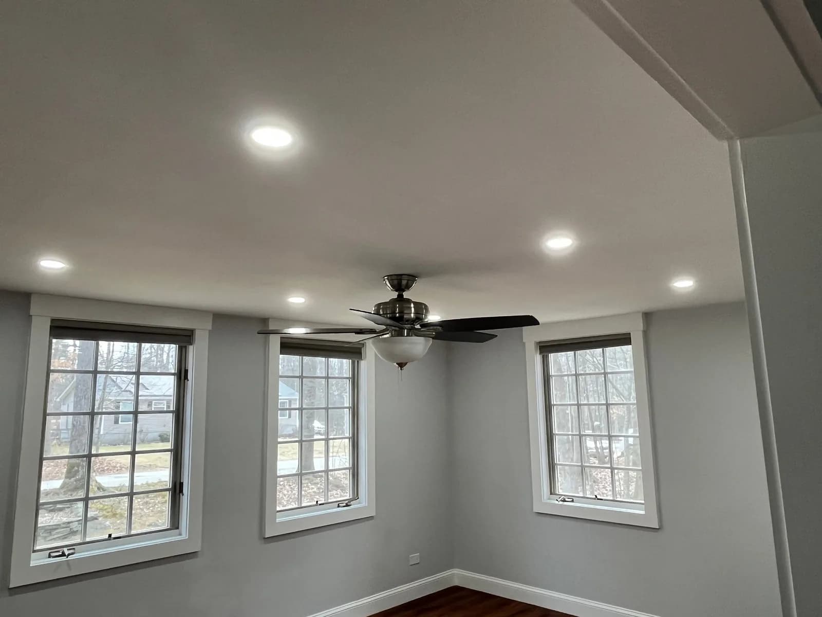 A freshly painted gray room with six lit recessed lights spaced across a smooth white ceiling around a brushed nickel ceiling fan, three white-trimmed windows looking out on a wooded street.
