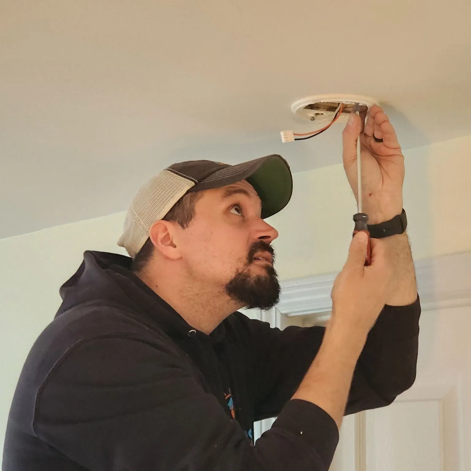 An electrician in a ball cap looks up while fastening a mounting plate with a wiring harness plug to the ceiling for a hardwired smoke alarm.