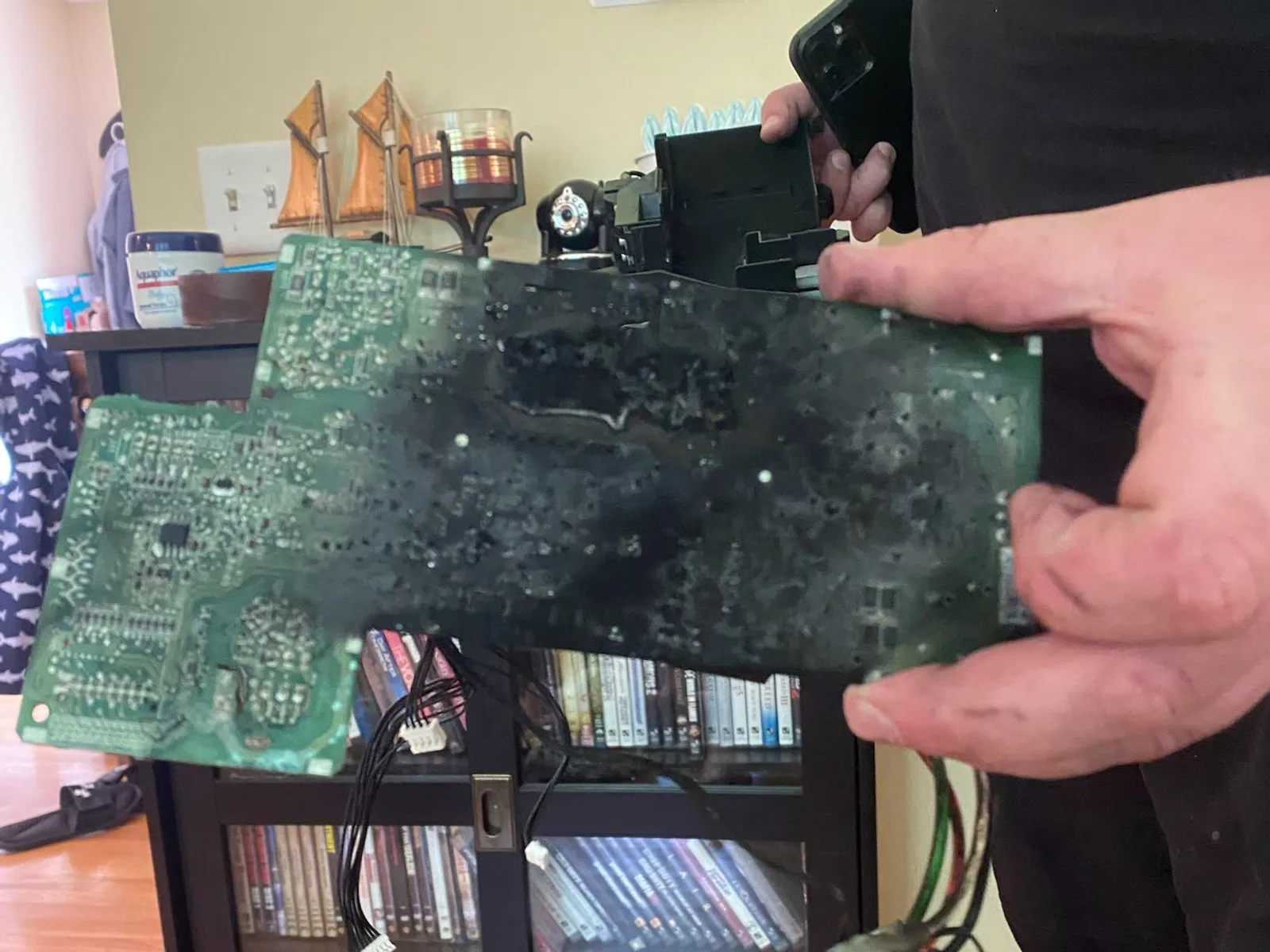 A hand holds up a scorched, blackened appliance circuit board with melted components in a living room, loose wires hanging from it.