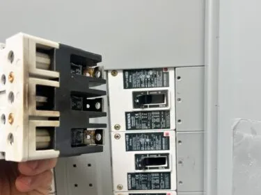 A hand holding a large multi-pole breaker beside an open Siemens load center filled with installed circuit breakers.