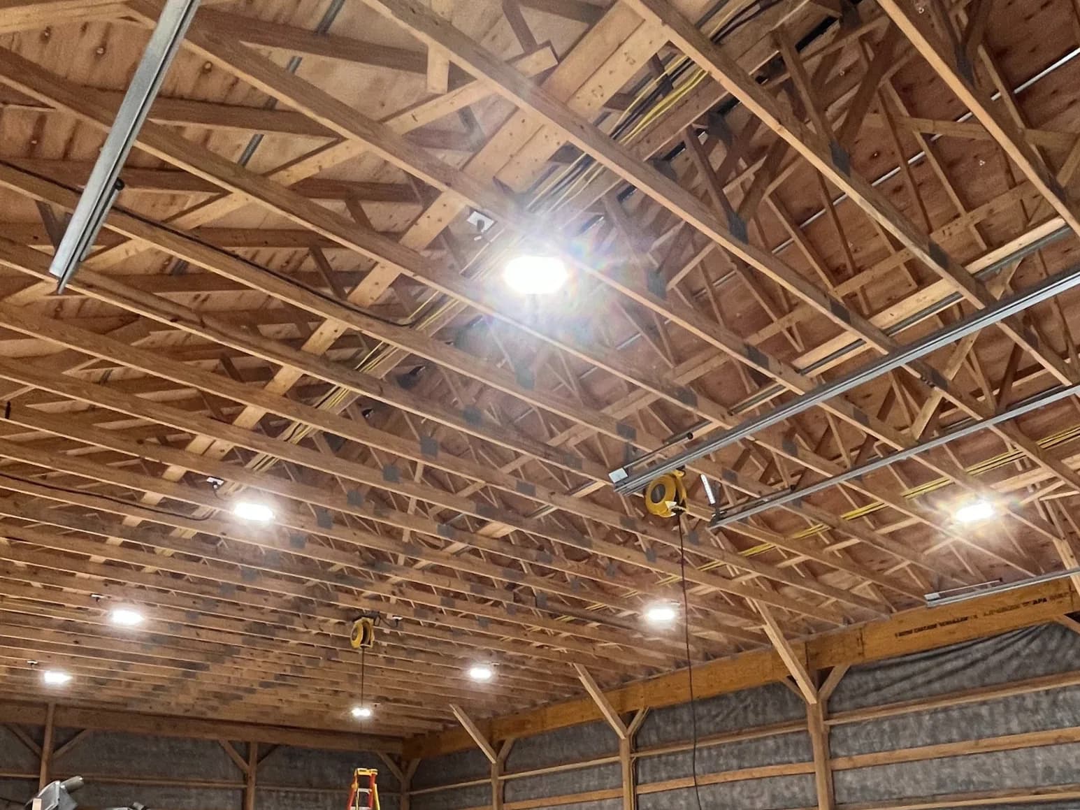 The interior of a large timber-framed pole barn with new high-bay lights illuminating the open wood-truss ceiling during a wiring job.