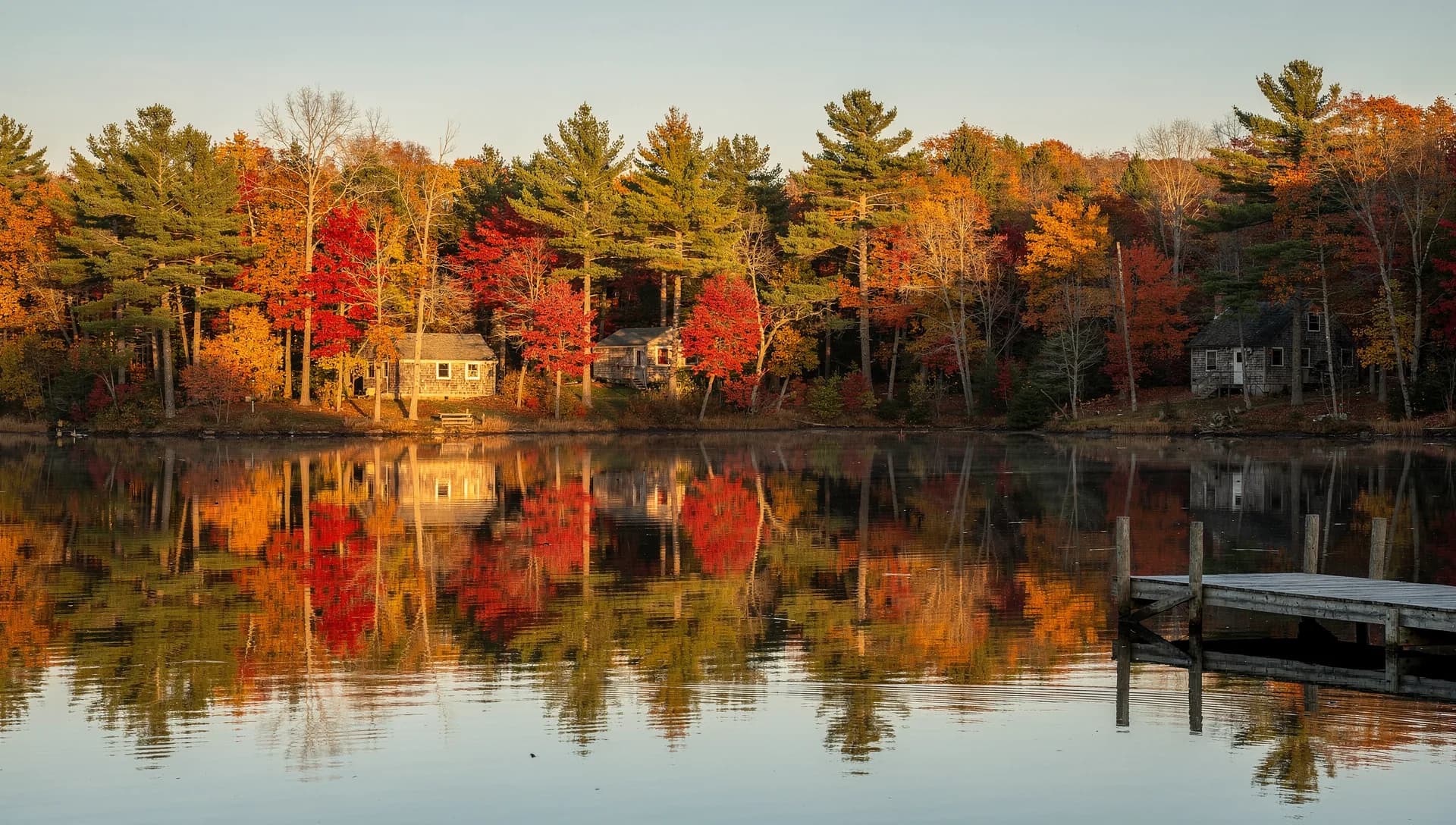 Shingled lakeside cottages among white pines and red maples, reflected in a still lake beside a wooden dock on an autumn evening in Barrington