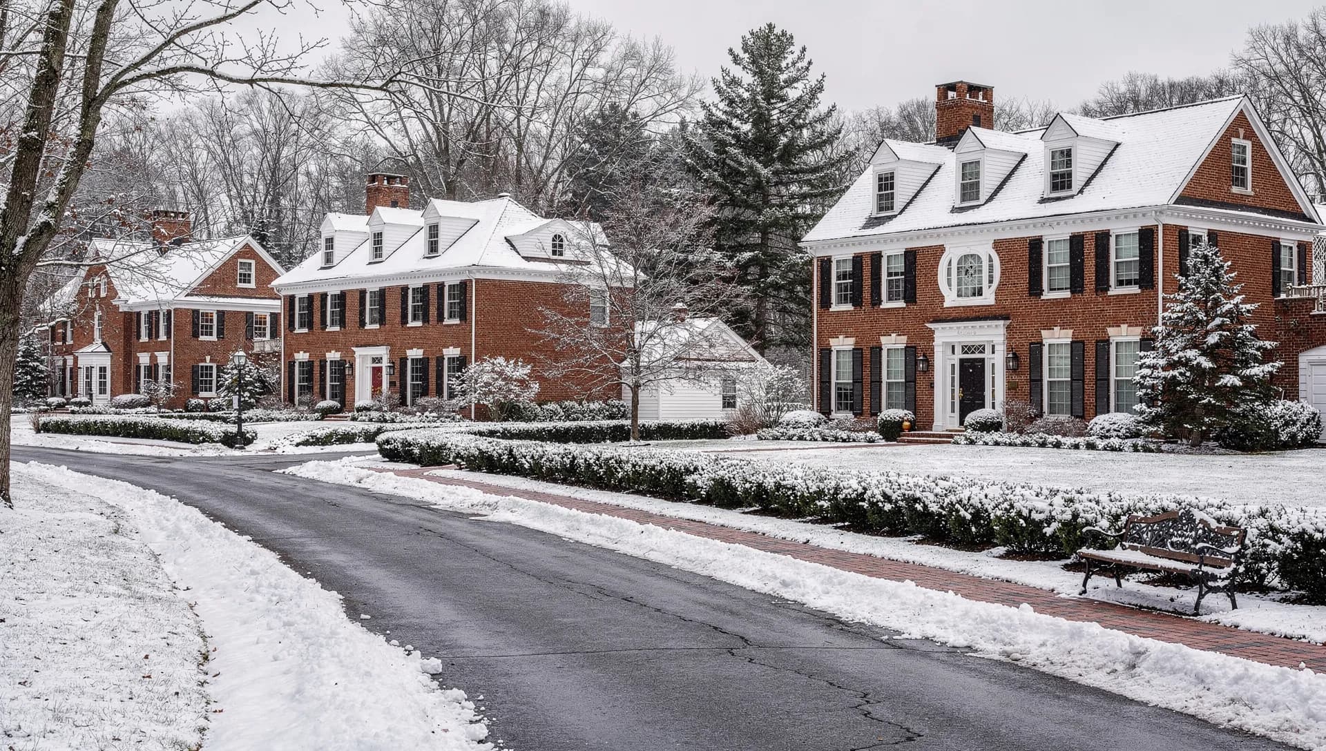 A street of large brick Colonial homes with black shutters and clipped hedges under a light fresh snowfall in Bedford, New Hampshire.
