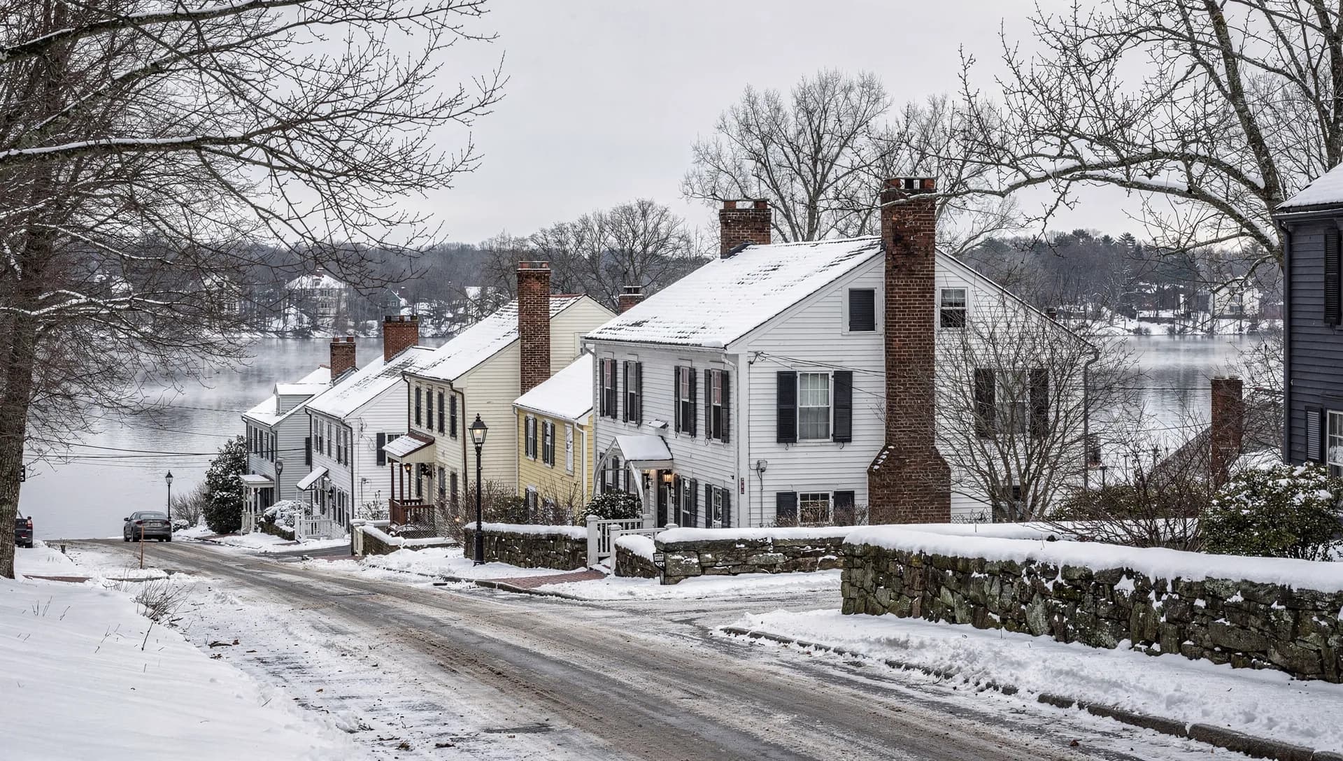 A row of white and pale-yellow colonial homes with brick chimneys along a snowy road above the Concord River in Billerica, Massachusetts.