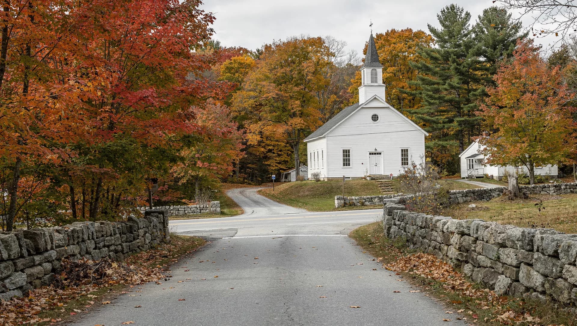 A white steepled meetinghouse on a rise above a stone-walled country crossroads framed by red and orange fall foliage in Danville, New Hampshire.
