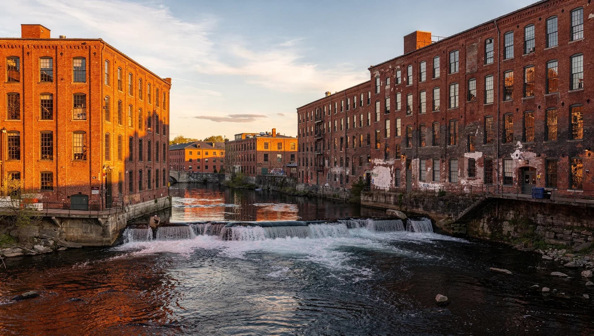 Red-brick mill buildings lining both banks of the Cocheco River above a low waterfall at golden hour in downtown Dover, New Hampshire.