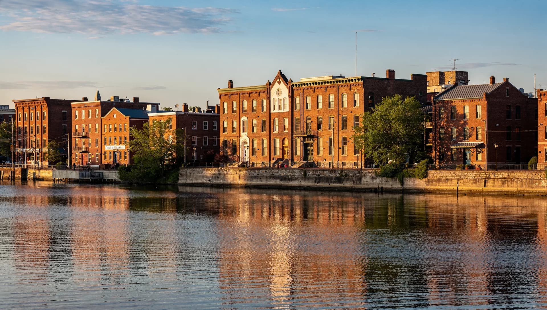 Tall red-brick mill and downtown buildings rising above a granite riverfront wall and reflected in the Merrimack River at golden hour in Haverhill, Massachusetts.
