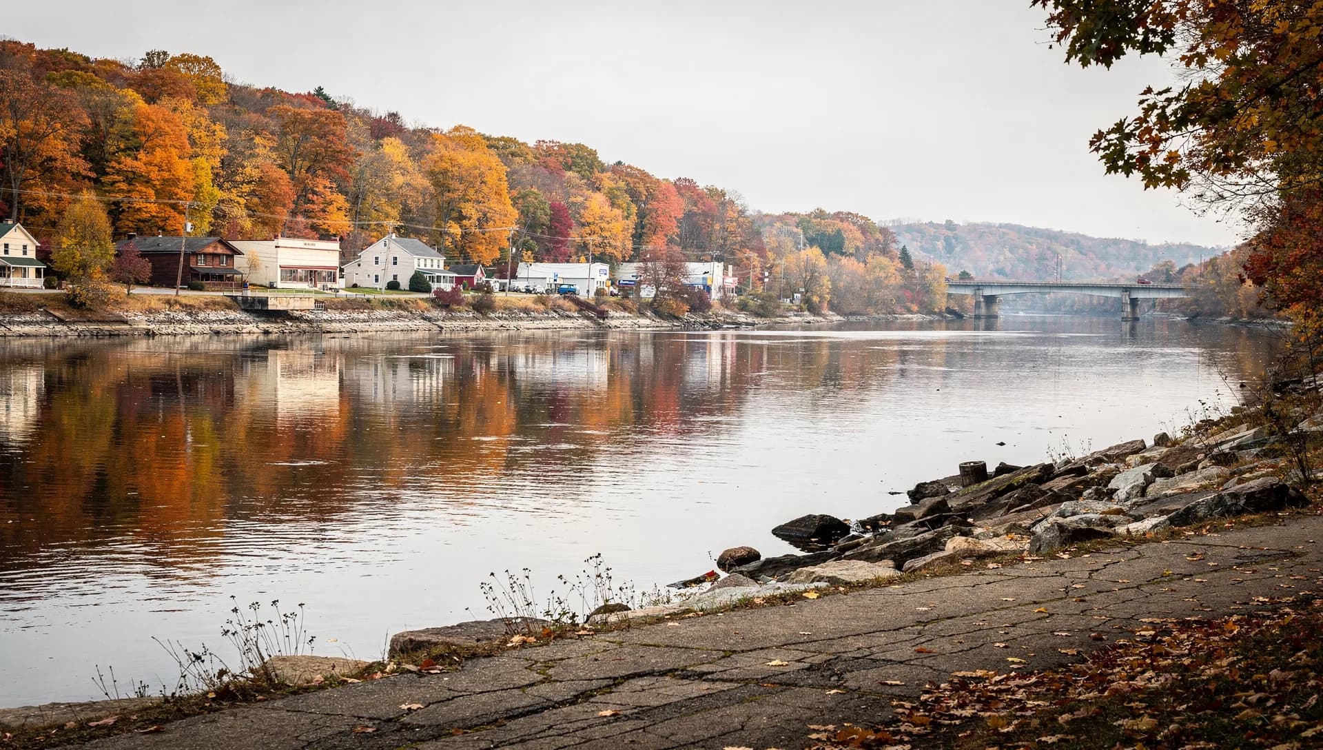 A wide, calm stretch of the Merrimack River reflecting autumn woods, with a small riverside village and a distant highway bridge, in Hooksett, New Hampshire.