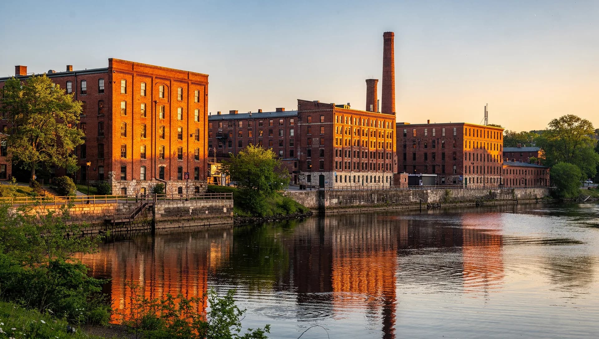 Historic red-brick textile mills lining a canal in downtown Lowell, Massachusetts, under a clear sky.