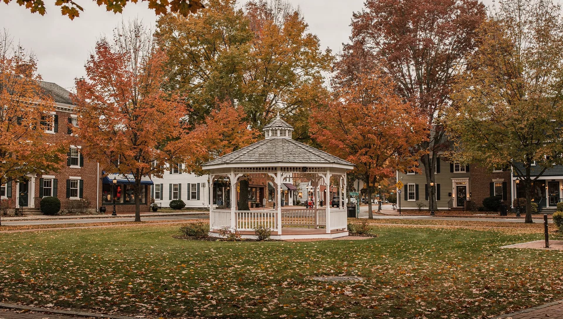 A white gazebo on the empty town green in Milford, New Hampshire, framed by autumn trees and brick buildings.