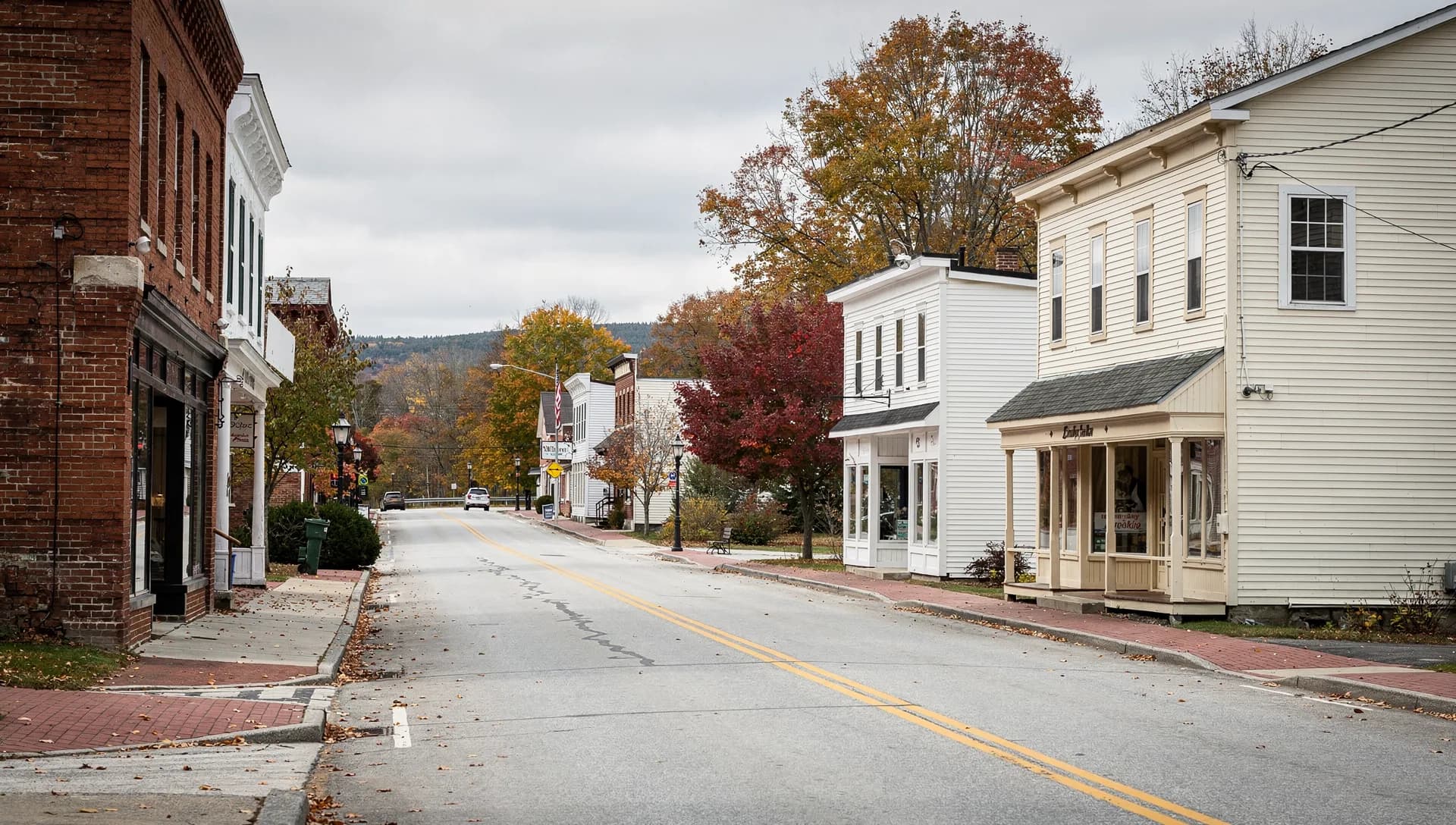 A quiet small-town main street of brick and white-clapboard storefronts under autumn trees, with wooded hills beyond, in Raymond, New Hampshire.