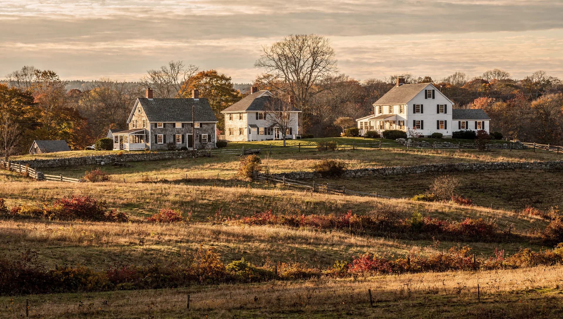 Rolling hilltop farm fields edged with stone walls and three white colonial farmhouses on the ridge at golden hour in Stratham, New Hampshire.
