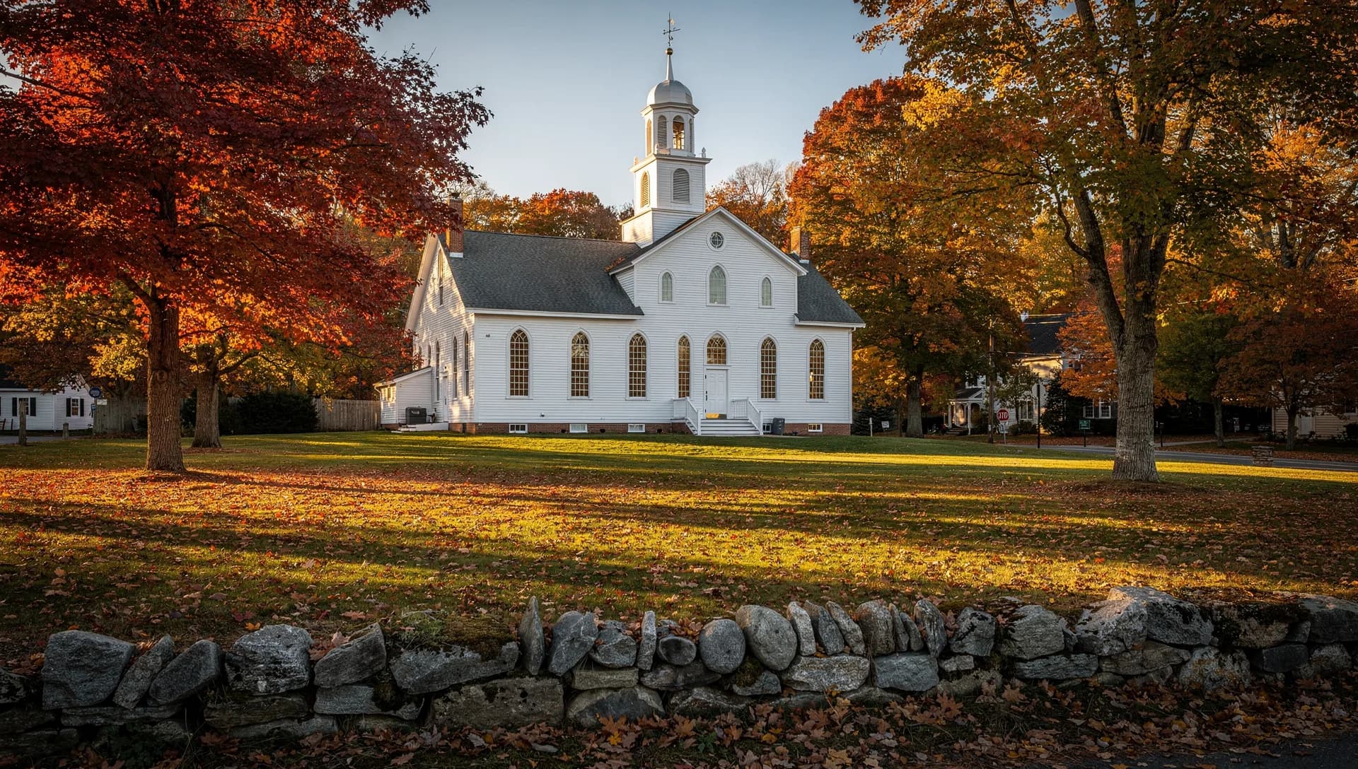 A white meetinghouse with arched windows and a tiered cupola on a sunlit green, framed by red maples behind a fieldstone wall in Wayland